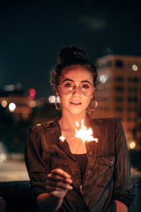 Mädchen mit Brille hält eine entzündete Wunderkerze. Im Hintergrund eine Stadt bei Nacht.